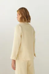 Chaqueta color crema de corte recto con cuello, solapa y bolsillos ojal. Forro solo en las mangas y terminaciones envivadas. Esquinas redondeadas y mangas con corte en puño que permiten transformarla en manga 3/4. Más larga en la espalda que en la delantera. Abrochadura con dos botones.