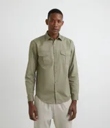 Camisa de hombre color verde oliva, de manga larga y corte clásico, confeccionada en sarga de algodón. Presenta cuello clásico, cierre frontal abotonado, dos bolsillos con solapa abotonada en el pecho y puños abotonados.