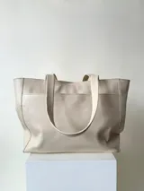 Cartera tipo tote color beige, hecha en cuero graneado con textura uniforme y suave al tacto. Tiene doble asa de hombro, un bolsillo frontal con broche imantado, un bolsillo trasero con cierre metálico y cierre principal también metálico. El interior está forrado y cuenta con tres bolsillos: uno con cierre, uno tipo plaqué para celular y un bolsillo grande tipo sobre con solapa y botón.