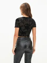 Top corto negro de manga corta, confeccionado en tejido de malla transparente con diseño de terciopelo floreado en relieve. Presenta cuello redondo y ruedo con terminación de ondas.