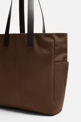 Bolso tipo shopper marrón de tejido de algodón con asas de cuero. Cuenta con dos bolsillos laterales exteriores y un bolsillo interno. Marca Mango.