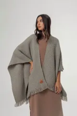 Ruana de punto color gris con diseño texturizado y terminación de flecos en el ruedo. Presenta un detalle de parche de cuero con logo en la parte frontal inferior.