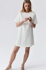 Vestido corto blanco de corte evasé, con cuello redondo y manga corta. Presenta abertura de cerradura con botón oculto en la nuca.