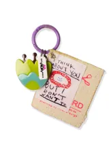 Llavero con aro metálico color lila, dije plástico con forma de flor verde y celeste, dije rectangular con la inscripción "SHE MONA" y dije de tela con la frase "I THINK ABOUT YOU BUT I DON'T WANT TO" y un dibujo de una cara sonriente.