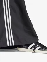 Pantalón deportivo negro de corte ancho con tres rayas blancas verticales en los laterales y logo de Adidas bordado en blanco en la parte superior de la pierna izquierda.