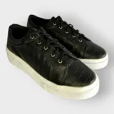 Zapatillas urbanas negras con textura de cocodrilo, cordones negros y suela de goma blanca.