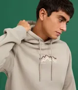 Hoodie cerrado de algodón con felpa, color beige claro, corte comfort. Presenta capucha con cordón ajustable, bolsillo canguro y puños y ruedo ajustados. Estampa minimalista de montañas en el pecho con líneas en tonos marrón, negro y rosado.