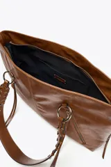 Bolso shopper de hombro color marrón, con flecos y trenzados en el cuerpo. Tiene doble cremallera metálica, asa doble con detalles metálicos y trenzados, y bolsillo interior. Cierre mediante cremallera metálica.