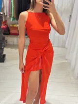 Vestido largo color naranja vibrante, sin mangas y con cuello redondo. Presenta un diseño drapeado y anudado en la cintura que genera una abertura alta en la falda.