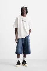 Camiseta blanca de corte relajado, cuello redondo y manga corta. Presenta un estampado de gráficos y textos combinados a contraste en la parte delantera y trasera.