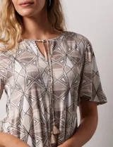 Blusa de tejido elastizado con estampado geométrico en tonos beige y blanco. Tiene cuello redondo con escote en V y lazo de ajuste, y mangas cortas amplias. Marca Cupio.