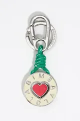 Llavero con mosquetón y anilla metálica plateada grabada con el logo de Bimba y Lola. Presenta un cordón trenzado verde con detalles en blanco que sujeta un dije circular con un corazón rojo esmaltado en el centro y el nombre de la marca calado alrededor.