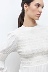 Blusa de color crudo con cuello alto fruncido y pechera con nido de abeja. Cuenta con mangas largas con detalles elásticos y puños con volados, y un bajo de corte peplum.