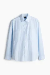 Camisa celeste a rayas verticales azules, de corte holgado, manga larga, cuello inglés, tapeta clásica, bolsillos superiores abiertos, canesú con pliegue de caja en la espalda y bajo levemente redondeado.
