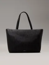 Bolso tote negro Calvin Klein, confeccionado en piel sintética acolchada con diseño de rayas verticales. Tiene doble asa de hombro, cierre superior con cremallera, herrajes plateados y logo metálico en el frente.