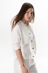 Chaqueta color crudo de lino con cuello redondo, manga tres cuartos, cierre frontal con botones y bolsillos de parche. Detalle de costuras en contraste color marrón.