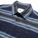 Camisa de franela a rayas horizontales en tonos azul marino, celeste y gris. Tiene cuello clásico, cierre frontal con botones, dos bolsillos de parche con botón en el pecho y mangas largas con puños abotonados.