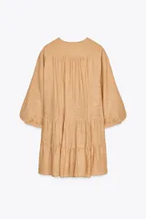 Vestido corto color beige con cuello redondo, cordón con borlas y manga larga. Presenta bordados combinados a contraste en la pechera, cierre frontal con botones ocultos, forro interior y bajo con volantes.