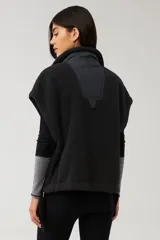 Chaleco de piel sintética tipo sherpa en color blanco crema, con cuello alto y cierre frontal mediante botones a presión. Presenta un diseño de hombros caídos y detalles de lazos decorativos en los laterales inferiores.