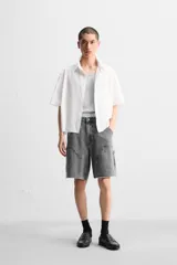 Bermuda color verde militar, de corte relaxed fit, con bolsillos frontales y traseros tipo plaqué, detalle de tira y bolsillo multifunción en la pierna, y bajo irregular.