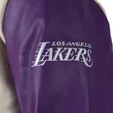 Campera bomber de algodón color crema con mangas de cuero sintético color violeta. Tiene el logo de Los Angeles Lakers bordado en el pecho y parches en los codos.