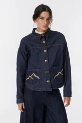 Camisa corta de jean azul con terminación redondeada en el ruedo. Presenta dos bolsillos plaqué bordados con motivo de montaña y detalle de pespunte a contratono.