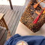 Bolso tipo tote bag con estampado animal print de leopardo en tonos marrones y negros. Tiene asas de cuero color marrón y un dije de borla roja.