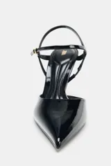 Zapato de tacón alto negro acharolado, con punta fina y diseño destalonado. Posee tiras finas ajustables con hebilla que rodean el tobillo.