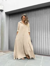 Vestido camisero largo con estampado de rayas verticales en beige y blanco. Presenta cuello camisero, cierre frontal con botones, mangas largas y tajos laterales en el ruedo.