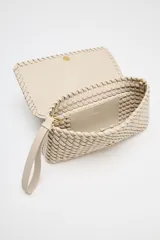 Bolso alargado color beige con diseño trenzado, asa bandolera y tirador con pulsera de mano. Incluye bolsillo interior y cierre mediante solapa y botón imantado.