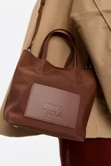 Bolso tote pequeño marca Bimba y Lola, modelo Chihuahua, color marrón. Confeccionado en nylon con estructura blanda, doble asa de mano y asa de tejido, piezas de piel, cierre con botón de presión, logo grabado, acabados metálicos, bolsillo interior e interior con placa metálica logotipada.