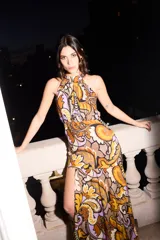 Vestido largo con estampado de arabescos en tonos marrón, naranja y lila. Tiene escote halter, corte ajustado en busto y cintura, falda fluida con tajo delantero y está confeccionado en seda de algodón con lurex.