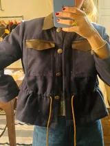 Chaqueta azul marino de paño con cuello y detalles en cuero marrón, cierre frontal con botones dorados, bolsillos con solapa y cordón ajustable en la cintura.