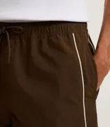 Bermuda deportiva masculina color marrón, con cintura elástica y cordón ajustable. Presenta piping contrastante en blanco a lo largo de los bordes y bolsillos funcionales. Confeccionada en poliamida de secado rápido.