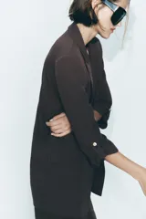 Blazer color marrón oscuro, abierta, con cuello solapa y hombreras. Manga tres cuartos con trabilla y botón dorado. Bolsillos delanteros con solapa.