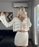 Conjunto de dos piezas tejido al crochet color crudo. Incluye una mini falda de tiro alto y un cardigan corto con mangas largas y lazos para atar en el frente.