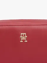 Cartera tipo cámara estructurada de color rojo, confeccionada en poliuretano. Presenta un cierre principal de cremallera y un monograma metálico TH en la parte delantera. Incluye una correa larga ajustable con detalles distintivos de la marca.