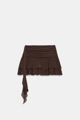 Falda pantalón negra de tiro bajo con cintura drapeada y bajo acabado en volantes superpuestos. Presenta un lazo largo y fino que cae desde el lateral.