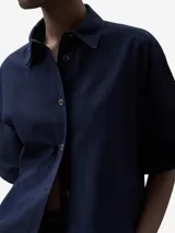Camisa azul marino de algodón seersucker con cuello solapa, manga corta sobre el codo, botonadura frontal y aberturas frontales en el bajo.