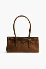 Cartera marrón de gamuza sintética con doble asa corta, cierre magnético y correa decorativa con hebilla metálica.