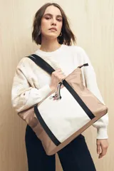 Bolso tipo tote de nylon color beige con panel central blanco y detalles de tiras negras. Tiene asas de hombro negras y cierre superior.