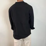 Camisa negra de manga larga con cuello mao y tapeta corta con botones de madera. El tejido presenta una textura arrugada o plisada. Las mangas están remangadas y sujetas con una presilla abotonada.