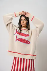 Sweater tejido color crudo con cuello redondo y mangas largas. Presenta dos peces rojos con detalles en azul bordados en el frente. Tiene ribete ondulado rojo en cuello, puños y ruedo.