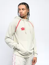 Canguro de felpa color crema con capucha, cordones y logo Umbro bordado en rojo. Presenta ribete rojo en hombros y bolsillo tipo canguro con cierre lateral.