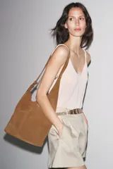 Bolso tipo saca asimétrica de serraje color marrón. Tiene asa bandolera ajustable y cierre con imán. Incluye cartera con bolsillo extraíble.
