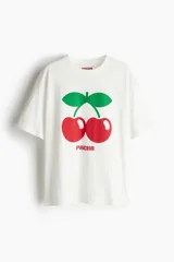 Remera blanca de algodón de corte amplio con cuello redondo y mangas cortas. Estampado frontal de dos cerezas rojas con hojas verdes y el logo de la marca Pacha en rojo.