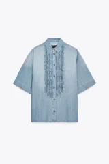 Camisa de denim celeste con cuello solapa y manga corta. Presenta detalle de volantes en el frente y bajo asimétrico con aberturas laterales.