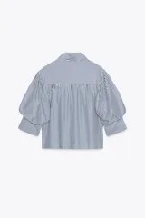Camisa de algodón a rayas verticales azules y blancas, con cuello solapa y mangas cortas abullonadas. Cierre frontal con botones.
