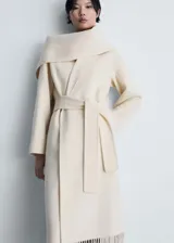 Abrigo largo color crema, con cuello con solapa, lazo a la cintura, bufanda desmontable y terminación de flecos.