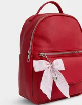 Mochila pequeña de cuero sintético color blanco con bolsillo frontal con solapa y cierre superior. Incluye un charm colgante de moño a rayas rojas y blancas con dije de corazón plateado.